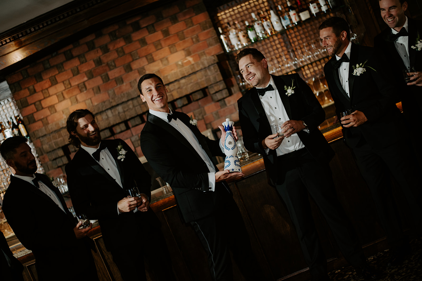 Smiling groom holding a tequila bottle while celebrating with his groomsmen at a classic bar.